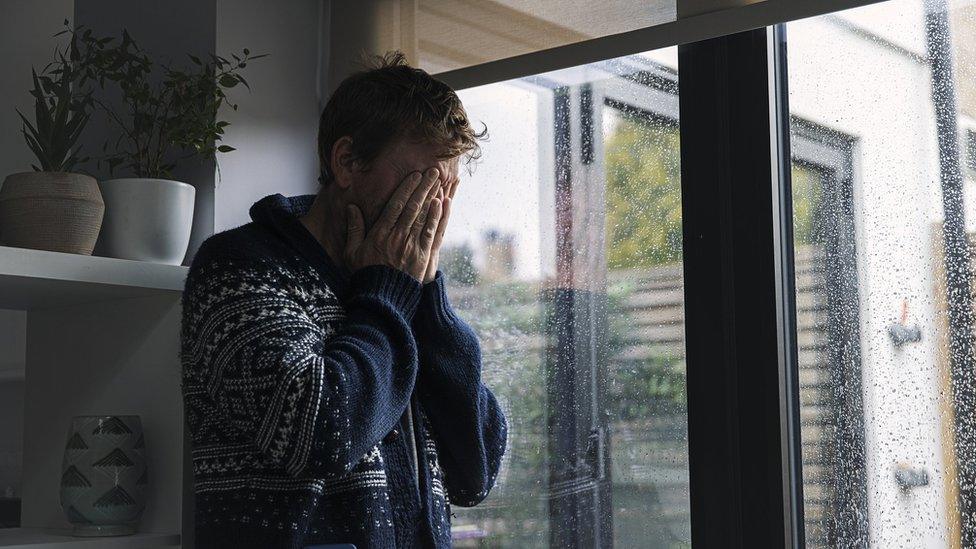 Man holding his hands to his face with rain on windows behind him