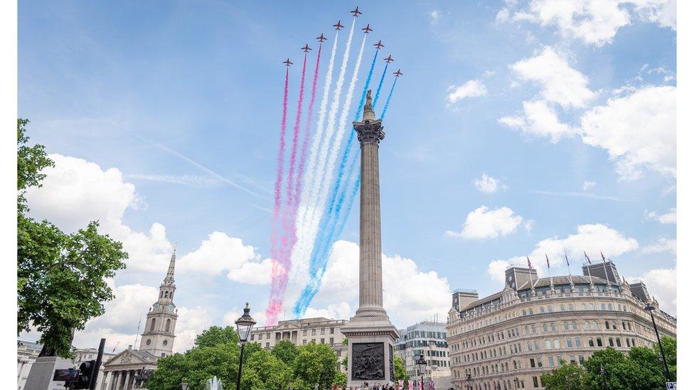 a group of nine planes fly in an arrow formation with red white and blue smoke trailing behind on a sunny day