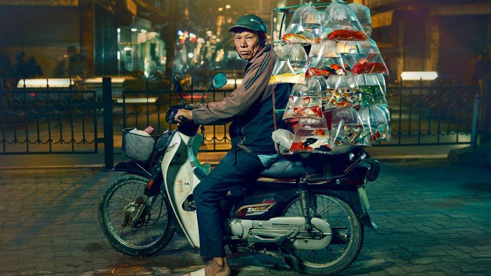 A man is posed on his motorcycle with an array of multicoloured fish in plastic bags hanging in rows on the back of the bike