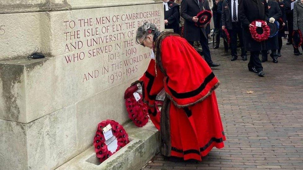 Woman in red cloak with black trim lays a wreath below the inscription on a war memorial