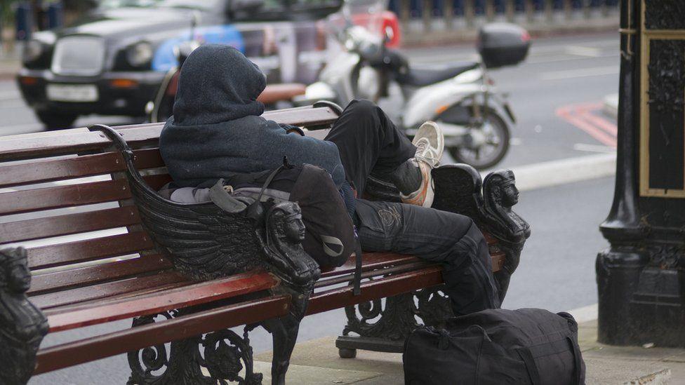 A rough sleeper wearing dark clothes facing away from the camera on a bench outside