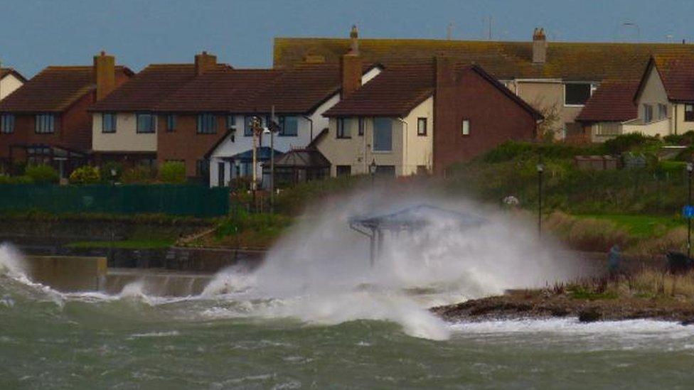 Waves and white foam shooting up onto the land in front of a cluster of houses