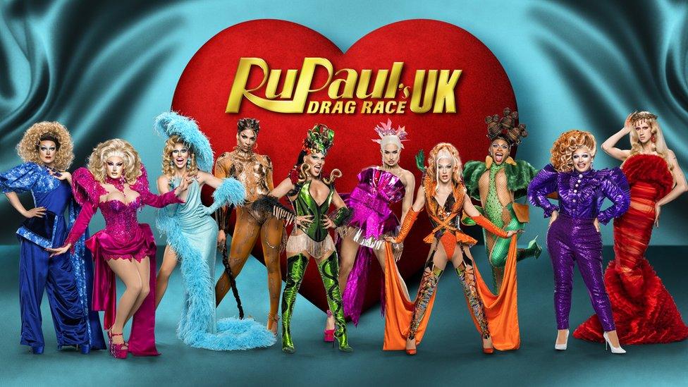 Promotional picture for the new series of Drag Race UK. The 10 drag queens are stood side by side posing with their hands on their hips or by their sides or on their heads. They're wearing a variety of brightly coloured dresses ranging from blue to pink to gold to green to orange to purple and red. The floor and background is light blue and behind them is a giant red heart with the words "RuPaul's drag race UK" in gold writing on it.