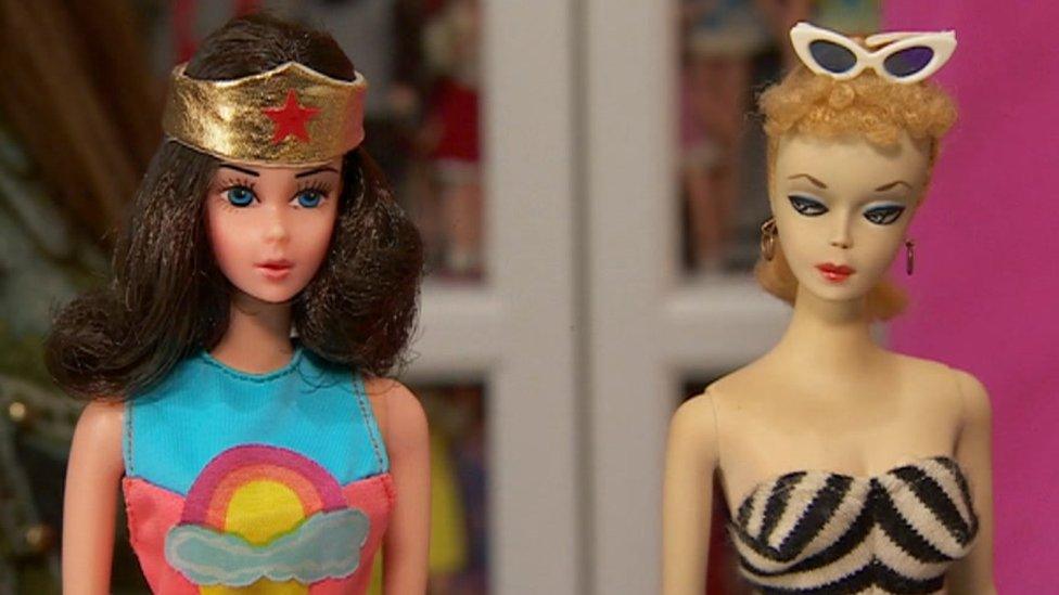 A close-up photograph of two Barbies, one wearing sunglasses and one wearing a superhero-style headband.