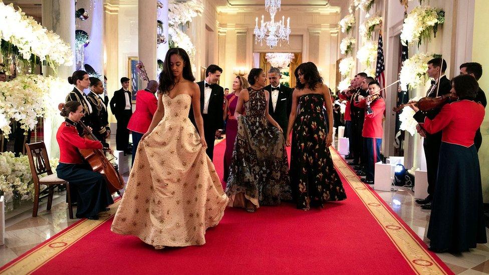 The Obama family, including the first lady Michelle and her two daughters, walk down a red-carpeted hallway flanked by musicians at a black tie reception in the White House.