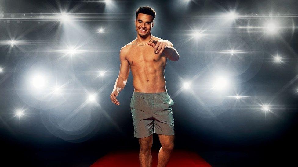 Toby, a shirtless man pointing at the camera wearing grey shorts. The background is of bright white spotlights on a black background