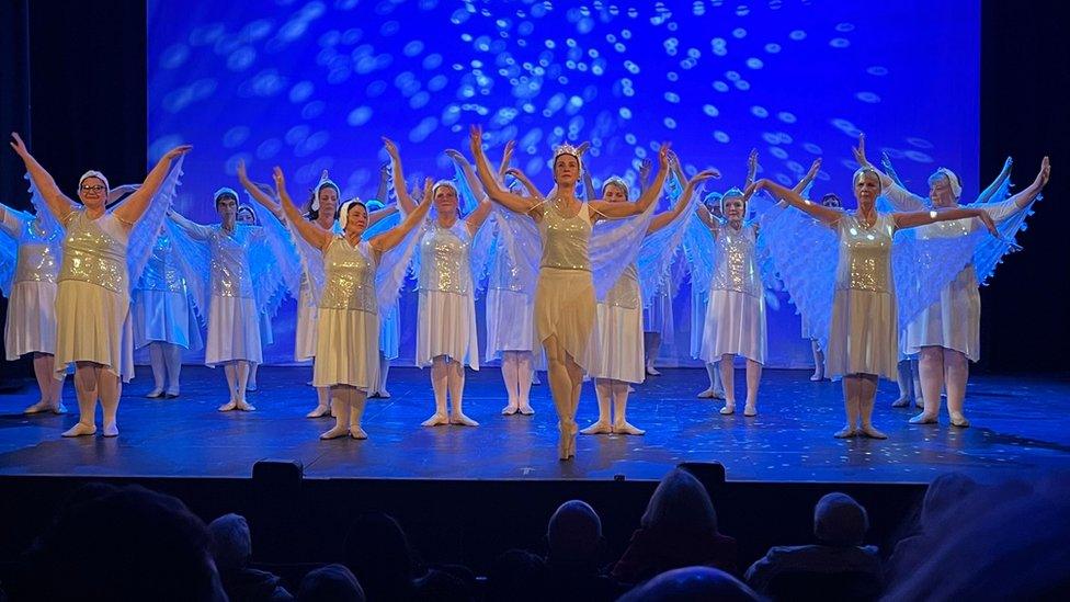 A group of women in silver and white costumes dance on a stage