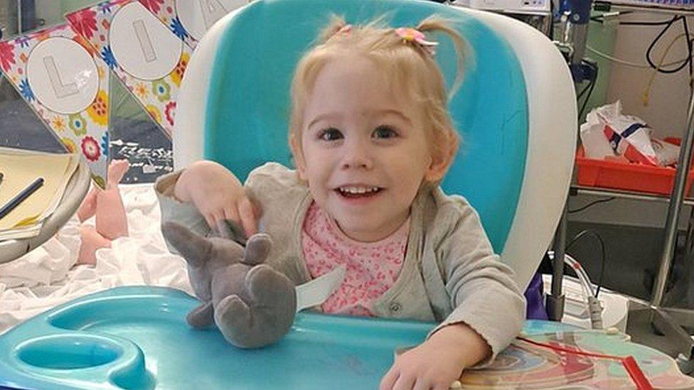 A little girl in a pink top sitting in a high chair holding a soft toy