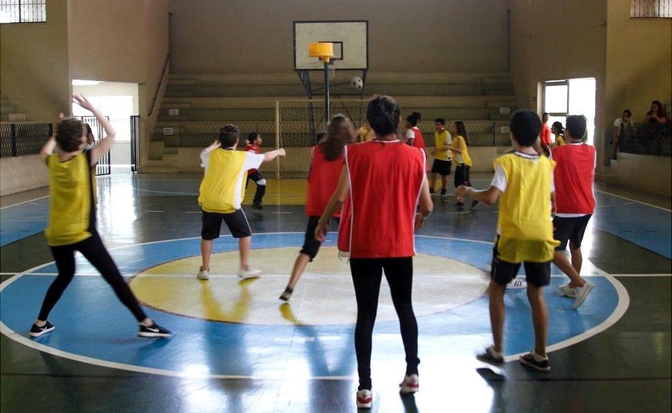 The children play Korfball, and a few of them have their hands in the air as a ball flies overhead near the yellow goal, high in the air