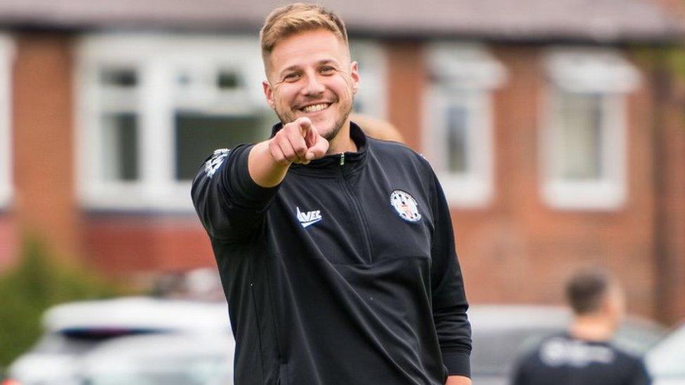 Adam Capper smiling for the camera with his right arm outstretched and his finger pointing directly at the lens. He is a white man with short blonde hair and a bit of stubble. He is wearing a black zipped up football jacket and behind him there are the windows of a building but they are blurred because it was taking on portrait mode.