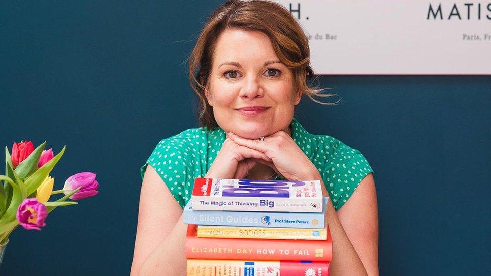 Louise Taylor in a green speckled blouse, smiling with her chin rested on her hands and peering over a pile of colourful books