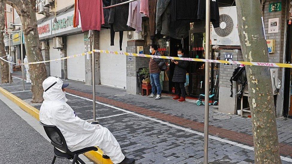 A police officer wearing personal protective equipment watching to two residents standing in front of their home