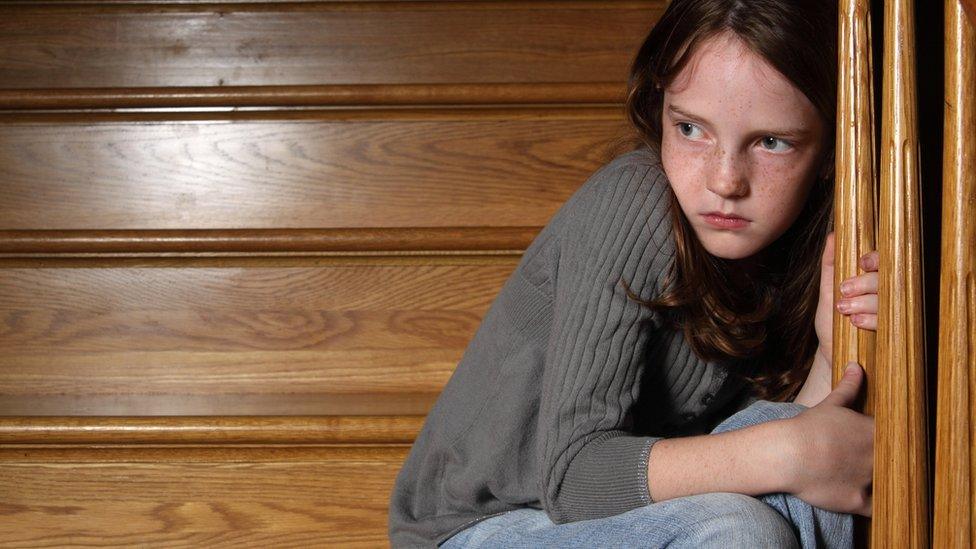 A young girl on a staircase looks frightened, gripping the bannister in fear