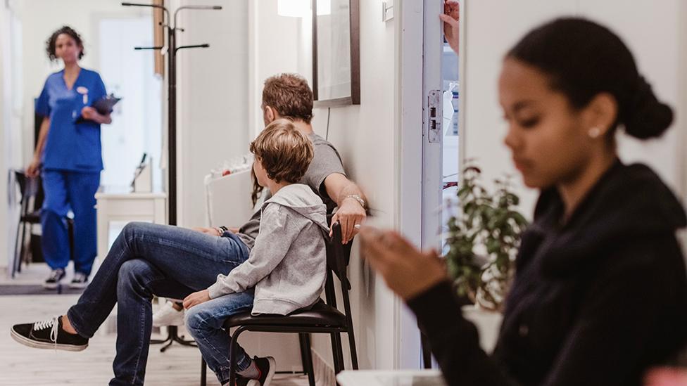 Patients wait at a hospital as a healthcare professional walks in the corridor