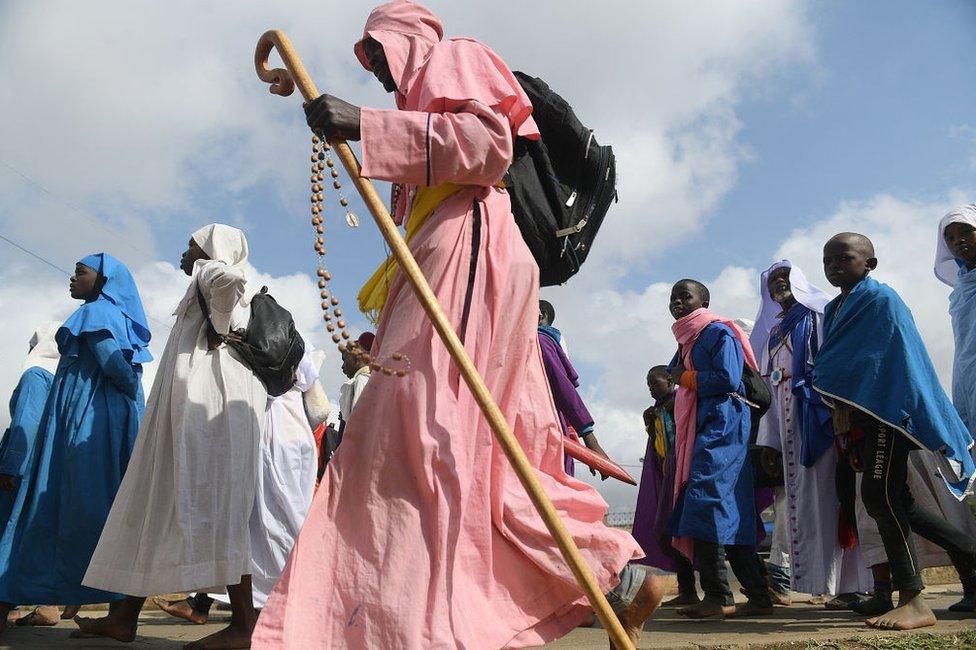 A man in a long, pink robe carries prayer beads and a walking staff. Others walking next to him are dressed similarly.