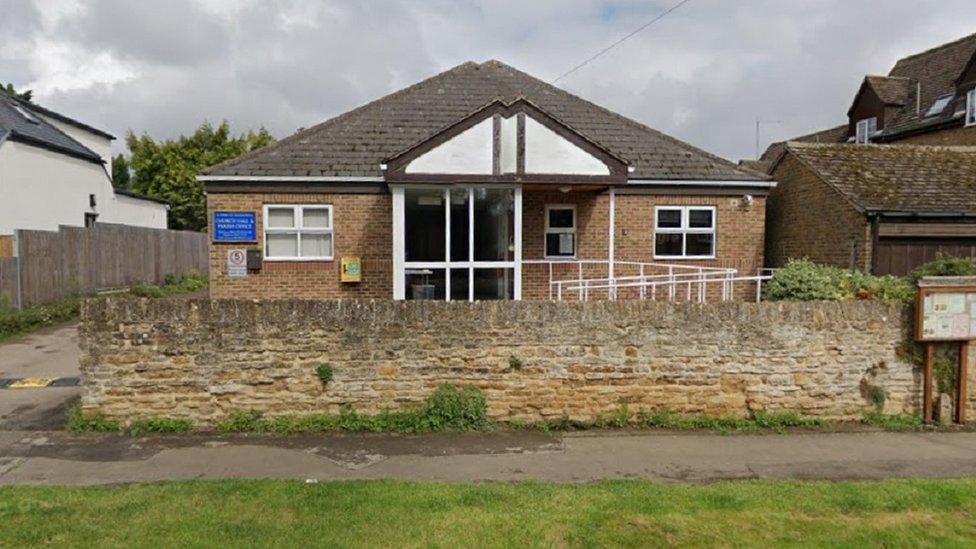 Single-story building with pitched roof and glass doors. Stone fence in front.