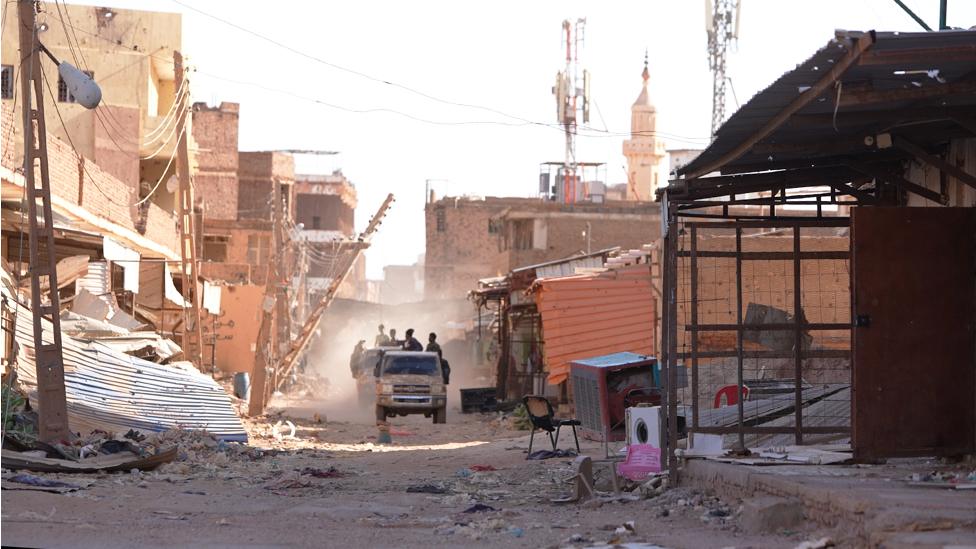 Army soldiers on back of pick-up truck driving through damaged street in Omdurman