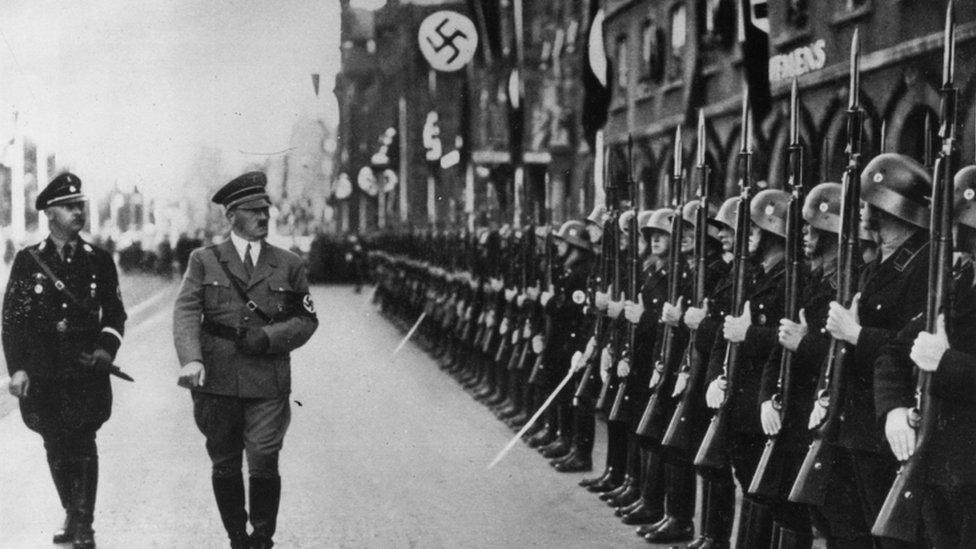 Himmler and Hitler walk by SS holding up large weapons in black and white image from 1938