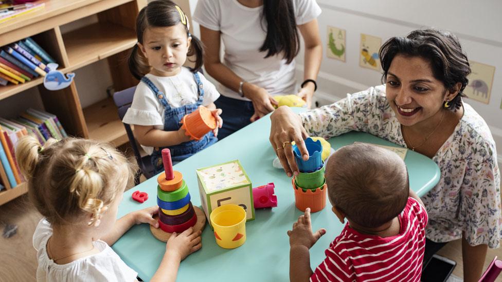 A woman playing with three young children around a table full of toys