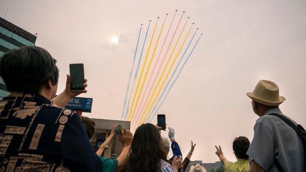 People in the streets of Beijing take photos and wave at planes flying in formation to Tiananmen Square