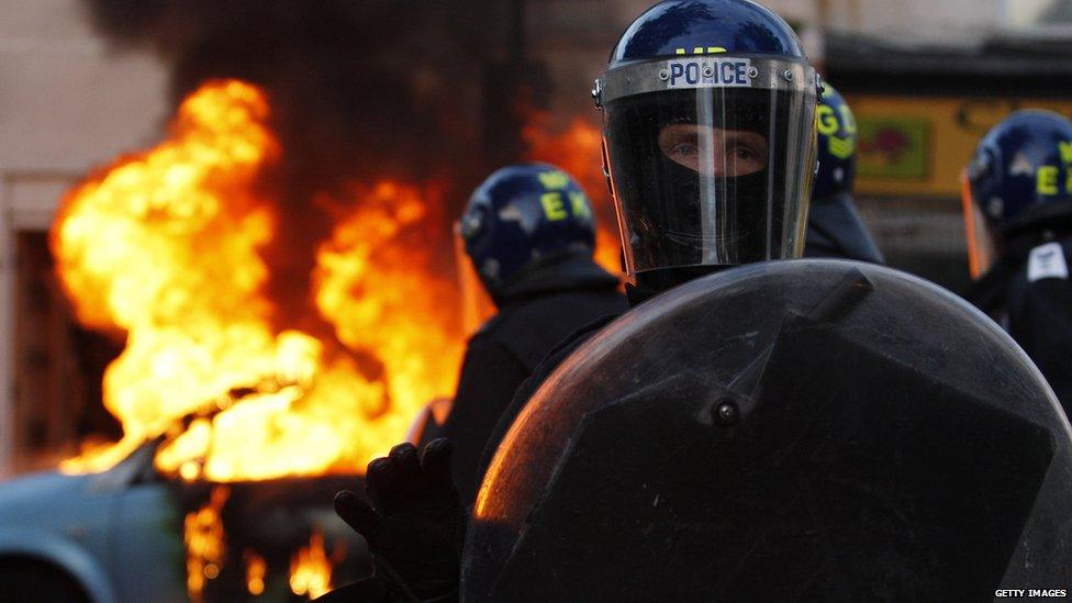 Riot police stand in front of a burning car during riots in London