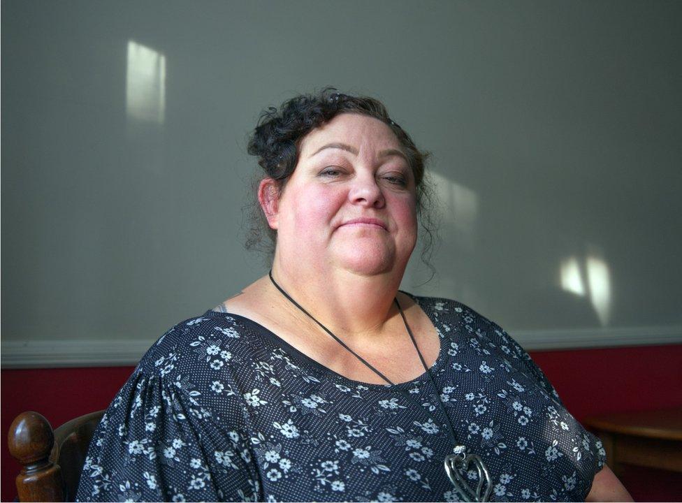 Helen Murphy, wearing a blue patterned dress, sitting at a wooden table. There are streaks of light coming in from the left of the frame which light up parts of the wall behind her.