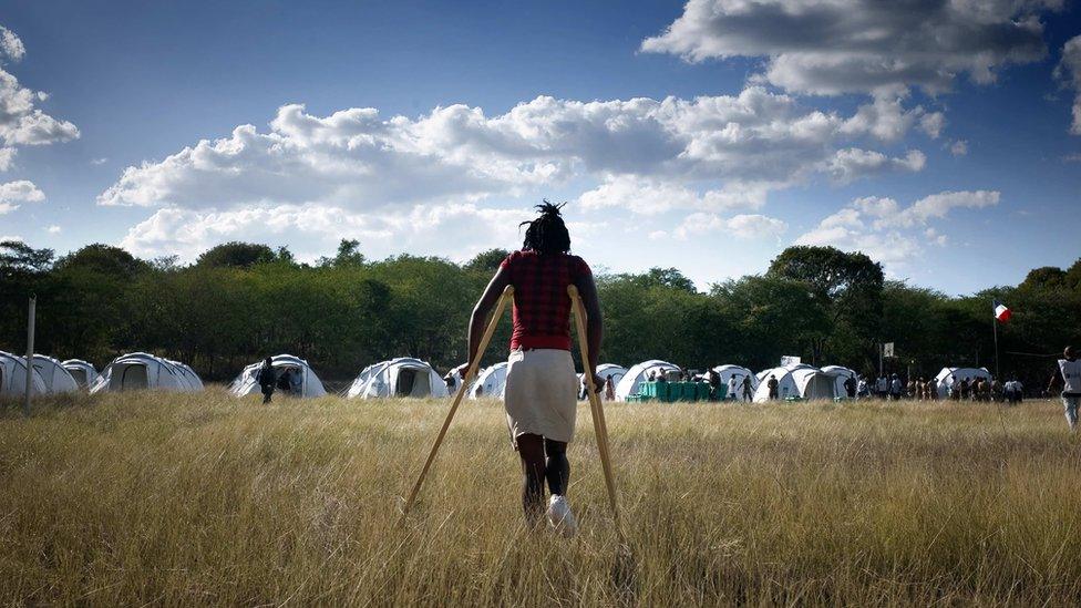 An injured young woman, on crutches, in front of a tent village being set up by charity workers / NGOs (in background), in Pandiassou, in January 2010.