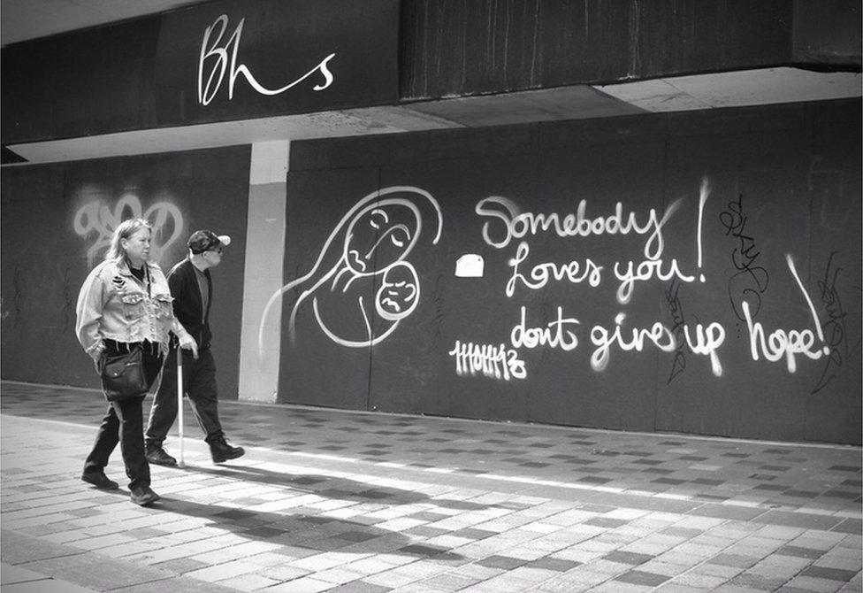 Two people walk down a high street and look at graffiti on a shop wall