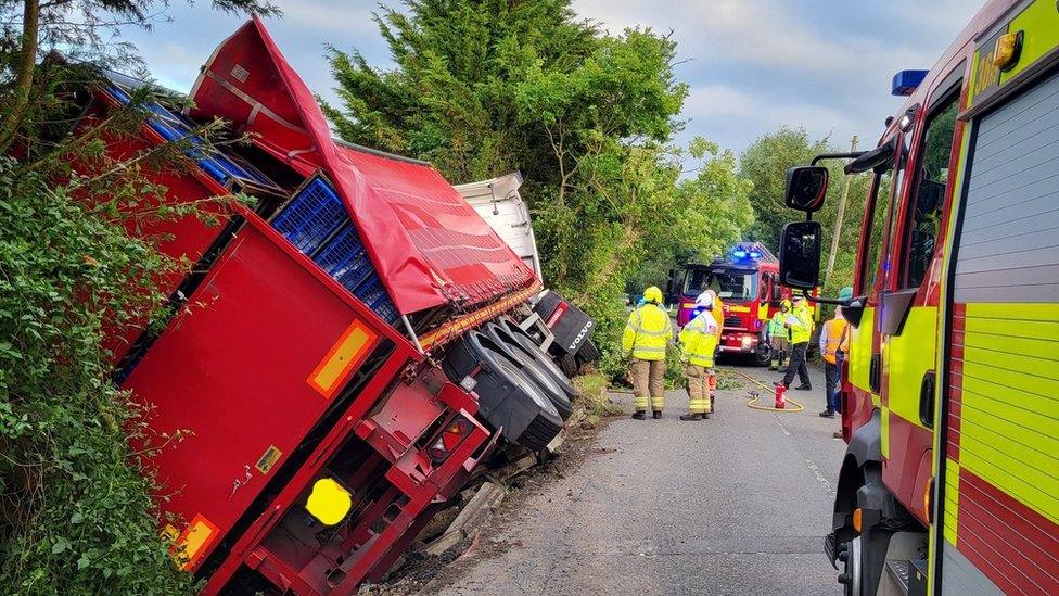 Lorry on its side, resting in hedging at side of road, with firefighters looking on