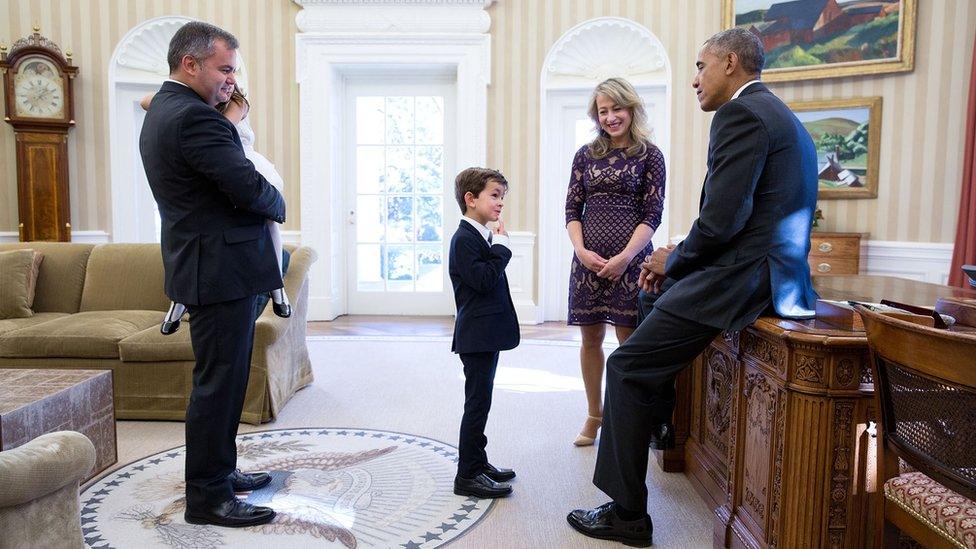 Obama, leaning casually against his desk, looks at six-year-old Alex, dressed in a suit, in a visit to the Oval Office. Alex's mother looks on as his father holds a smaller girl in his arms.