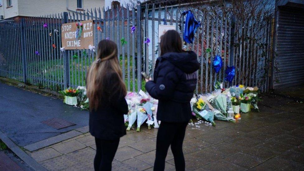 Photo of two girls from behind, stood next to flowers near to the scene in Knowle West where two teenage boys, aged 15 and 16, died after a stabbing attack.