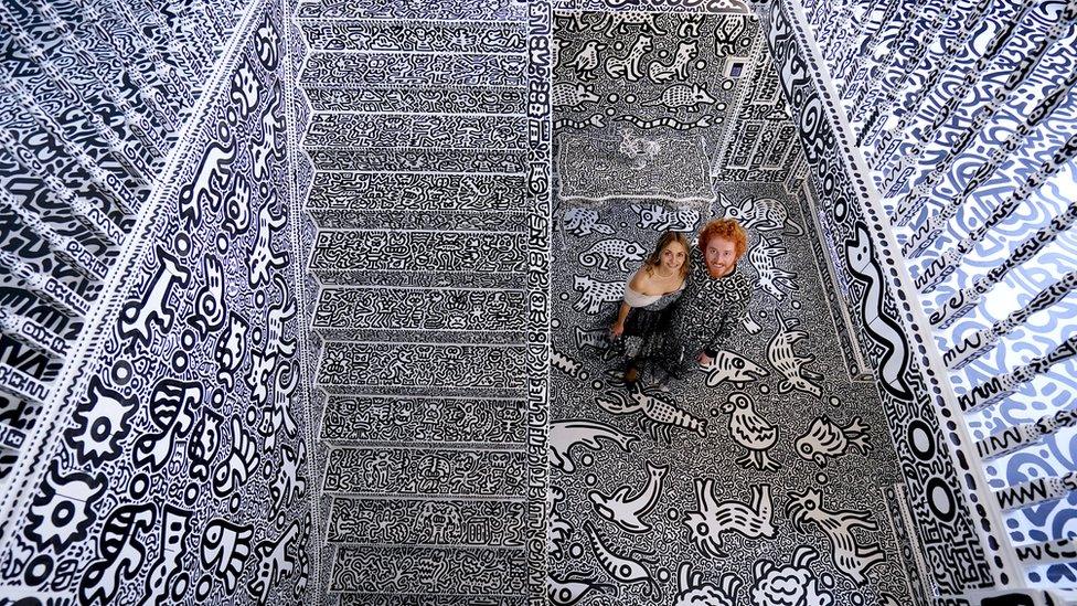 sam cox and his wife alena standing in their hallway at the bottom of the stairs looking up at the camera in the balcony above with every inch of the space covered in doodles