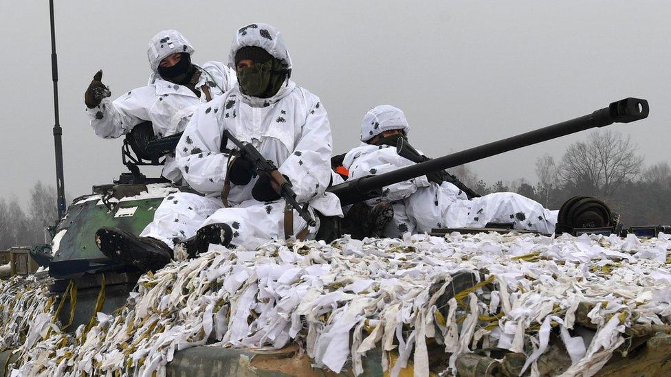 Three heavily armed Ukrainian servicemen clad in white camouflage site on top of a tank covered in similarly white fabric strips, with the tank's cannon protruding from the gap between the men