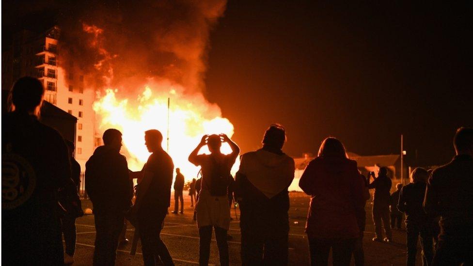 People celebrating the eleventh night are shown as silhouettes with the bonfire and apartment building visible in the foreground