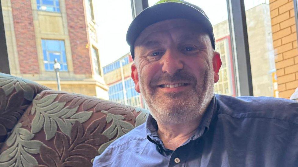 Stephen Black, a white man in a blue shirt and a black cap, smiles into the camera.