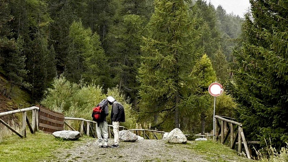 Two young men stand on a mountain gravel track on the Italian side of the French-Italian border, wearing trainers and carrying a small bag