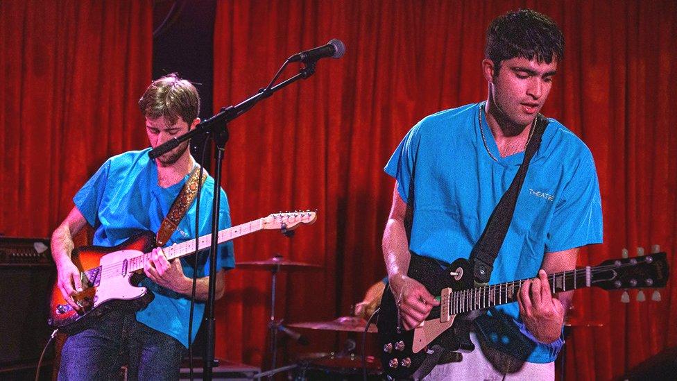 Two men stand on a stage in front of a red curtain, playing guitars. The man nearest the viewer wears a blue hospital scrub top with the worth "theatre" printed in gold block capitals on the left breast. He's got his instrument on a strap slung over his shoulder and is looking down as he forms a chord with his left hand and strums with his right. His band mate wears the same top and pulls a similar pose as he plays along. His face is slightly obscured by a mic stand and microphone in the middle of the stage.