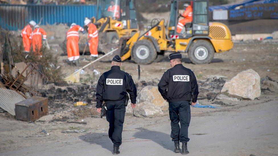 Two police officers walk towards a demolition crew on the site of the former camp at Calais