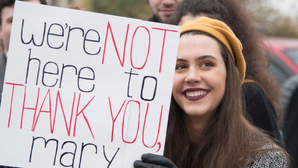 Woman holds up sign that says "We're not here to thank you Mary"