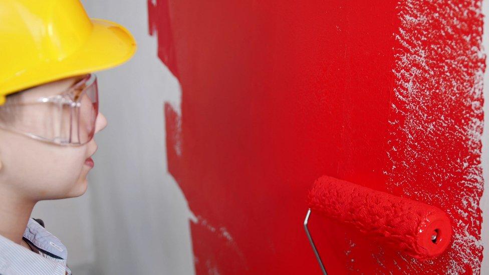 Boy wearing yellow hard hat rolls red paint on a wall