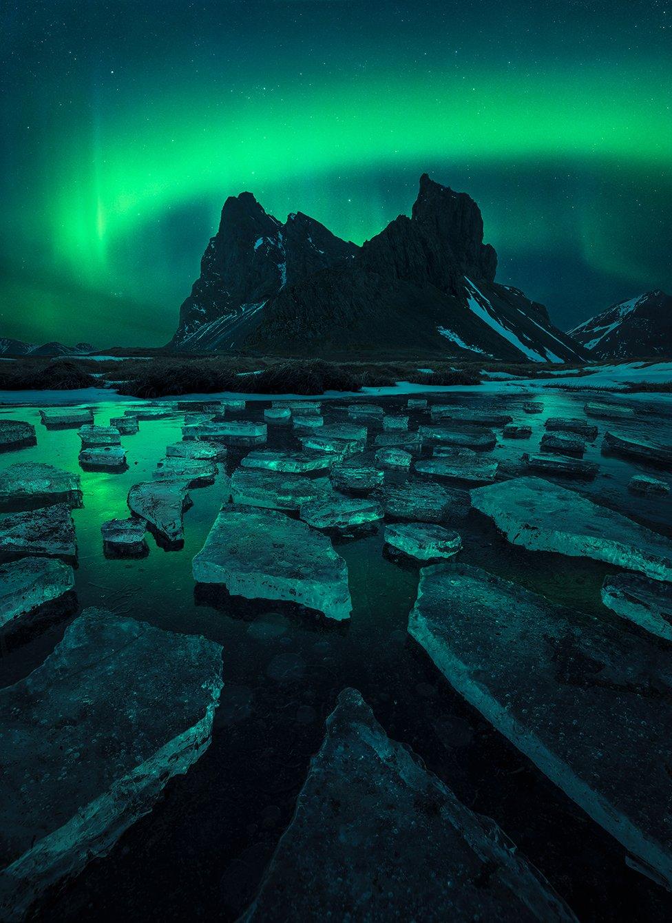 Mountain with northern lights in the background, and shards of ice on water in front.