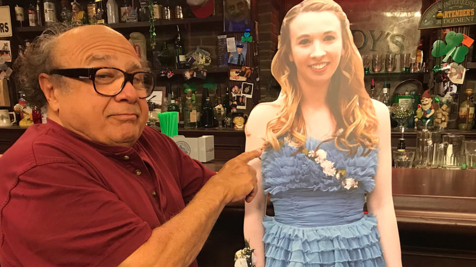 Danny DeVito stands next to a cardboard cutout of a teenager in a prom dress in Paddy's Pub, on the set of TV show It's Always Sunny in Philadelphia