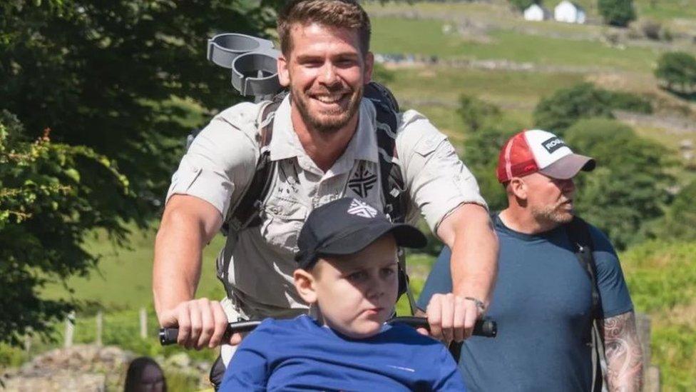 Man with short brown hair and beard pushes boy wearing blue cap in a wheelchair. Welsh scenery visible in the background.