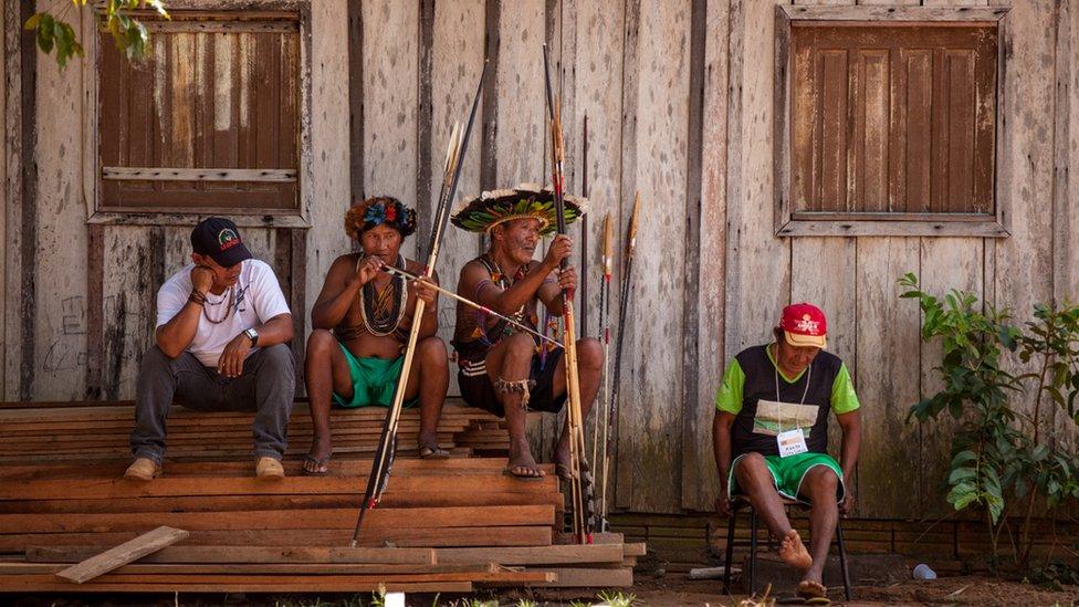 Members of the Cinta Larga sit by the side of a house with bows and arrows