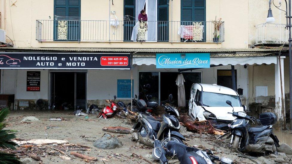 A woman looks down from a balcony at a street covered in debris and damaged vehicles