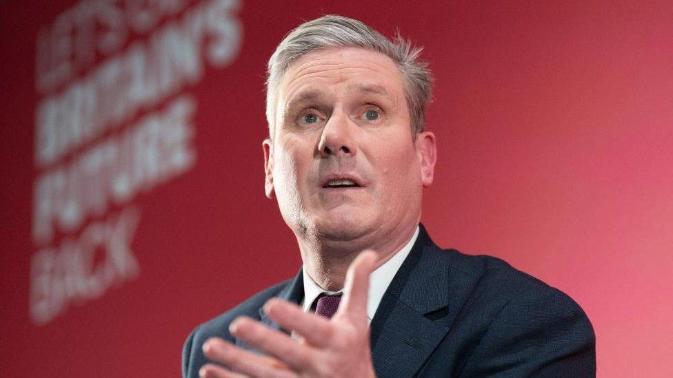 Labour leader Sir Keir Starmer seen given a speech on stage. The wall that forms the backdrop of the stage is red.