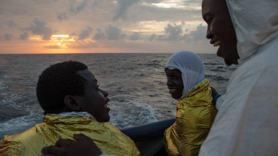 Three men wearing ponchos and survival blankets talk on a boat while sun sets in the background, the Mediterranean - Wednesday 23 November 2016