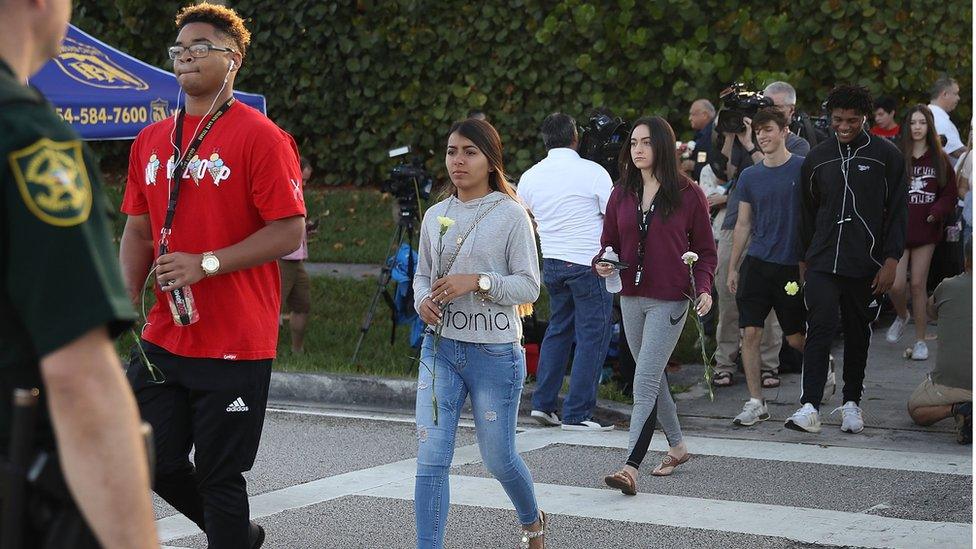 Students walk across a road holding flowers as they arrive at Marjory Stoneman Douglas high school