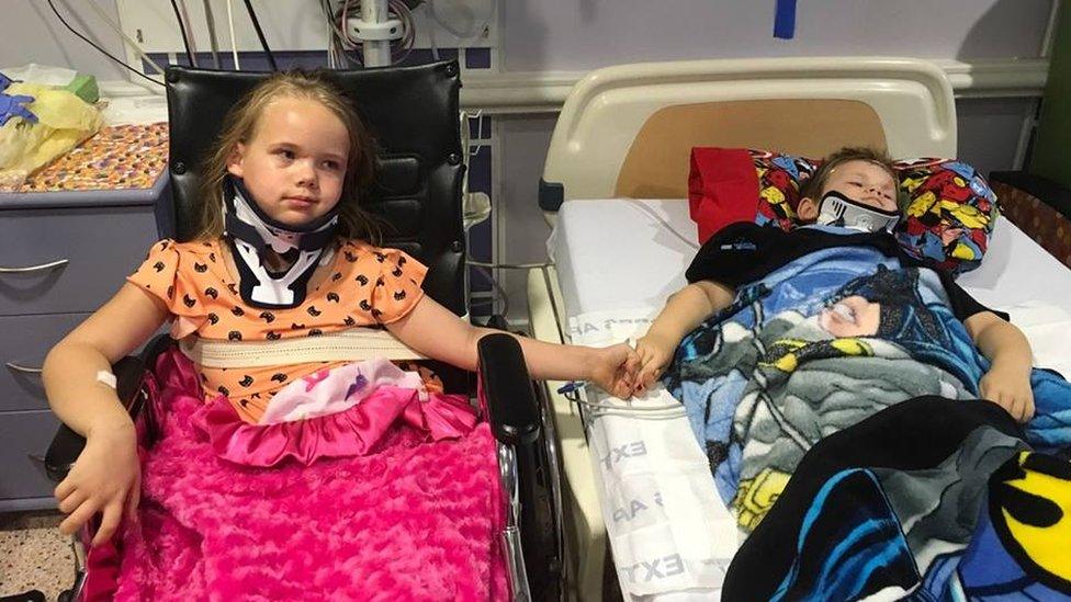 On the left, a young child sits in a wheelchair. She is holding hands with a boy lying in bed. They both have braces and are in a hospital.