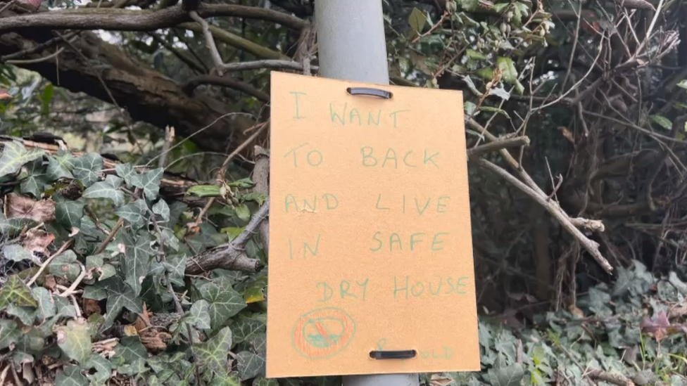 Child's handwritten poster saying "I want to back and live in safe dry house"